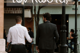 Two men in business attire walking away on a city sidewalk, seen from behind with storefronts and lampposts in the background.