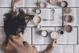 Overhead view of a person in a brown sweater lying among open books arranged in a grid, with numerous coffee mugs and spoons resting on the pages.